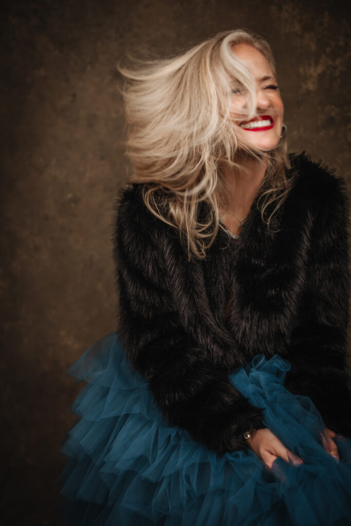 A woman with long blonde hair wearing a black fur jacket and blue tulle skirt smiles with her hair blowing across her face against a neutral background. Tanya Eivin Photography