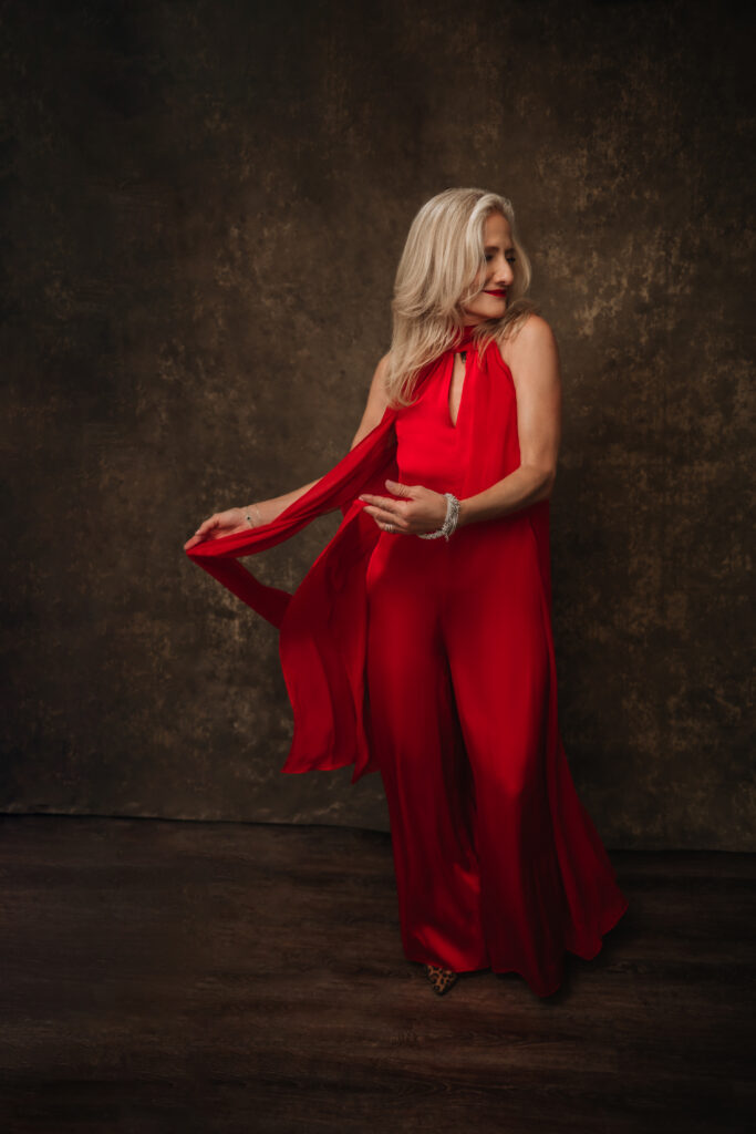 A woman with long blonde hair wearing a flowing red jumpsuit stands against a dark textured background, looking to the side and holding part of her outfit. Tanya Eivin Photography