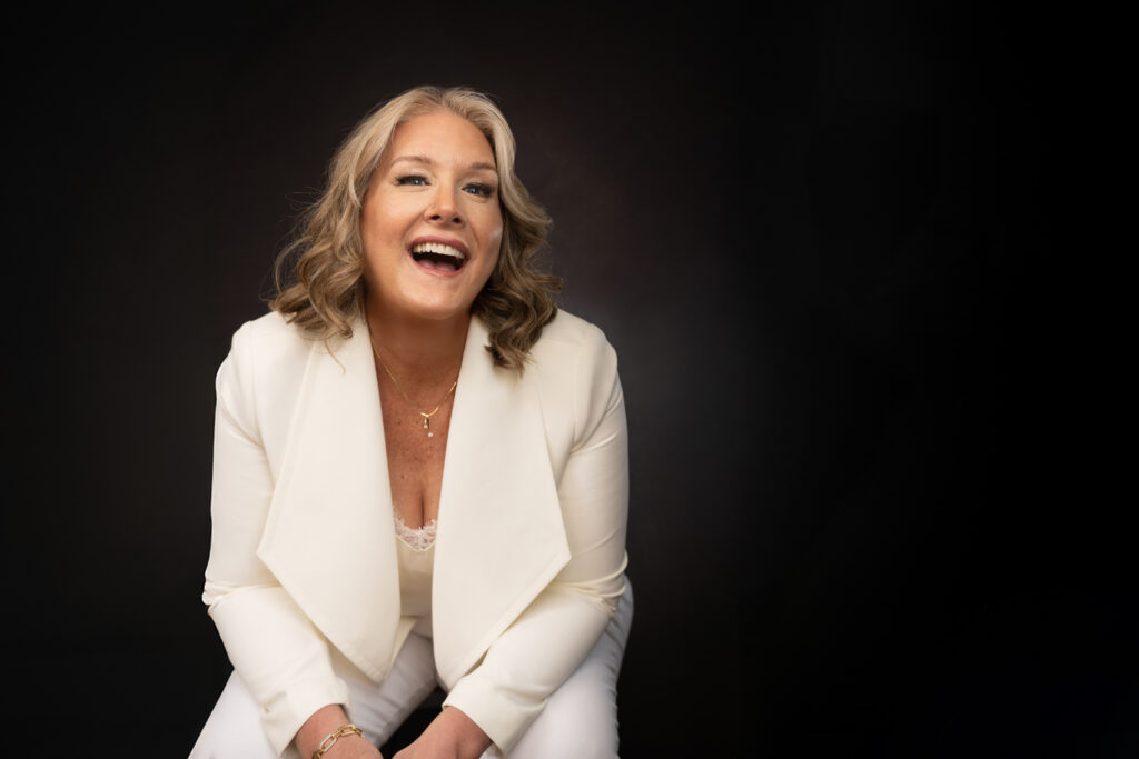 A woman with shoulder-length blonde hair wearing a white suit sits and smiles against a black background. Tanya Eivin Photography