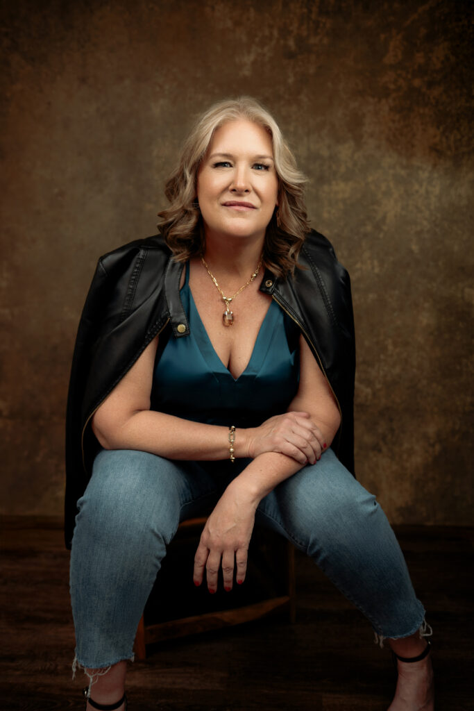 A woman with shoulder-length blonde hair sits on a stool, wearing a teal top, ripped jeans, and a black jacket draped over her shoulders, against a textured brown background. Tanya Eivin Photography