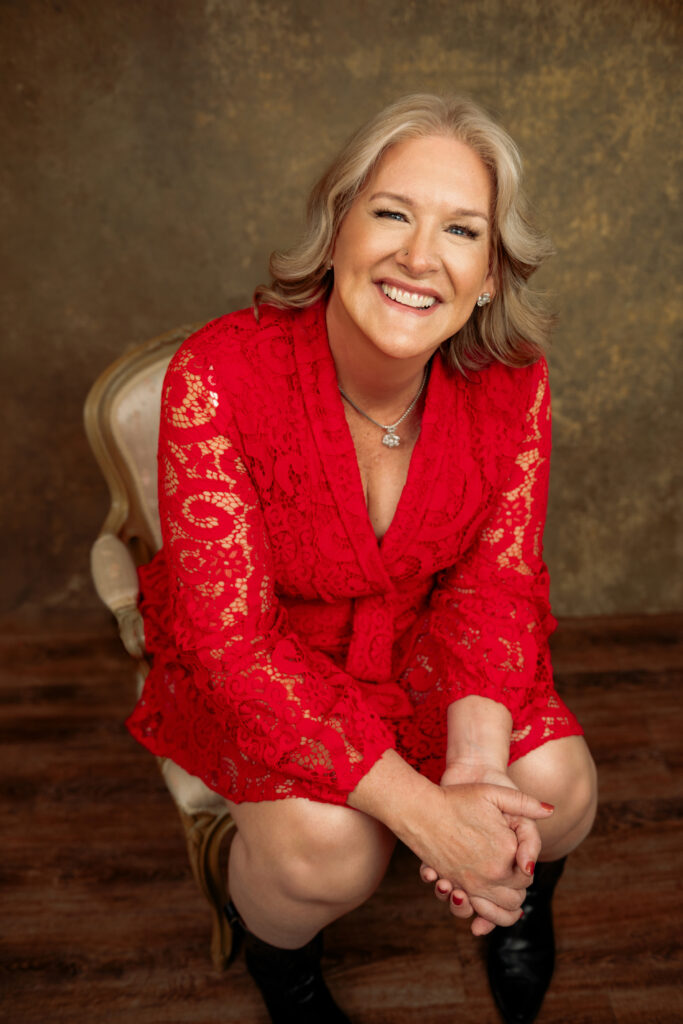 A woman with light hair in a red lace dress sits on a chair, smiling and looking up, against a brown textured background. Tanya Eivin Photography