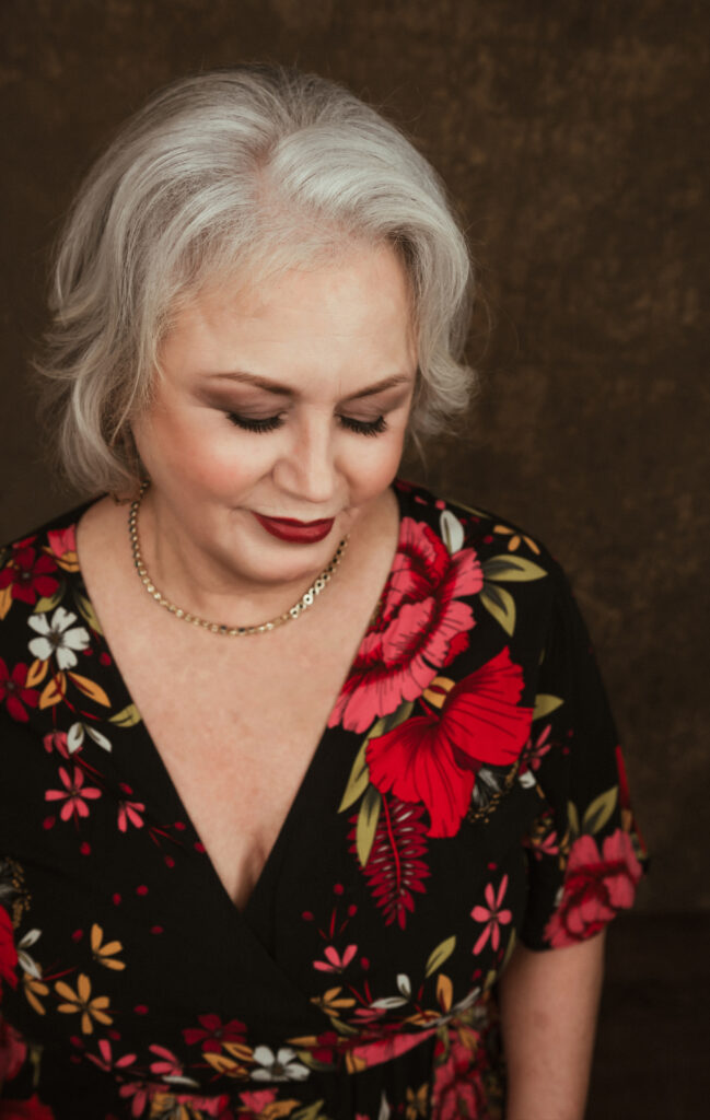Older woman with short gray hair and red lipstick, wearing a black floral dress and gold necklace, looks down with eyes closed against a brown background. Tanya Eivin Photography