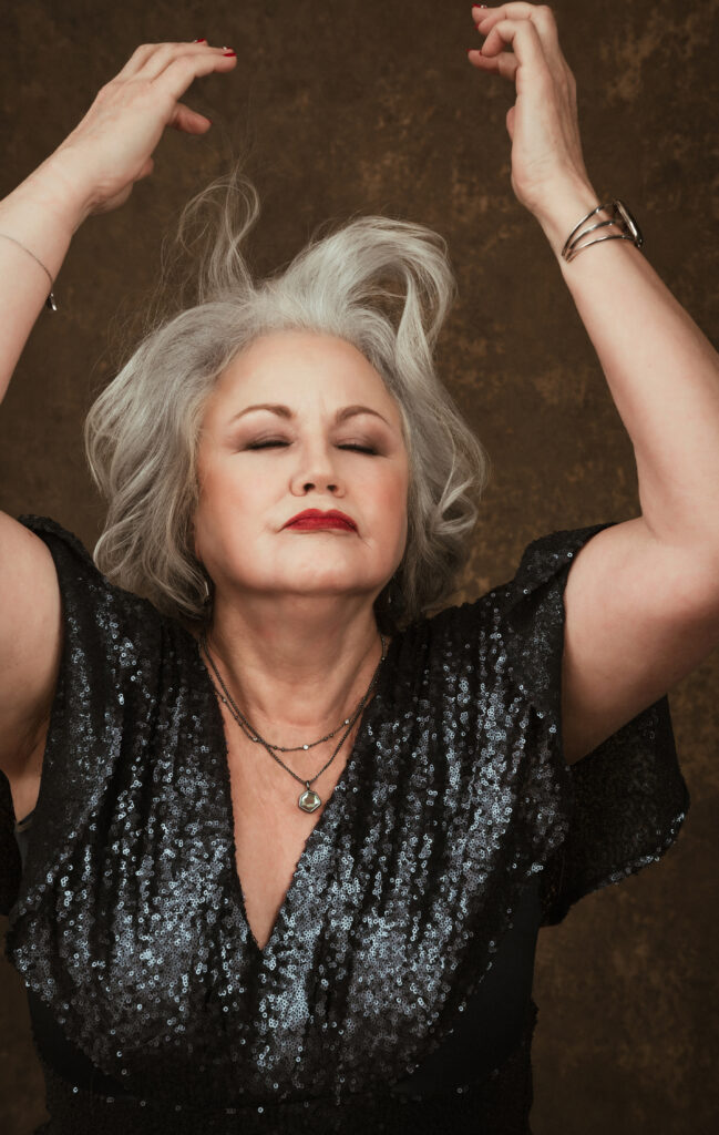 Older woman with gray hair, eyes closed, and arms raised, wearing a sparkly black top and layered necklaces, against a brown background. Tanya Eivin Photography