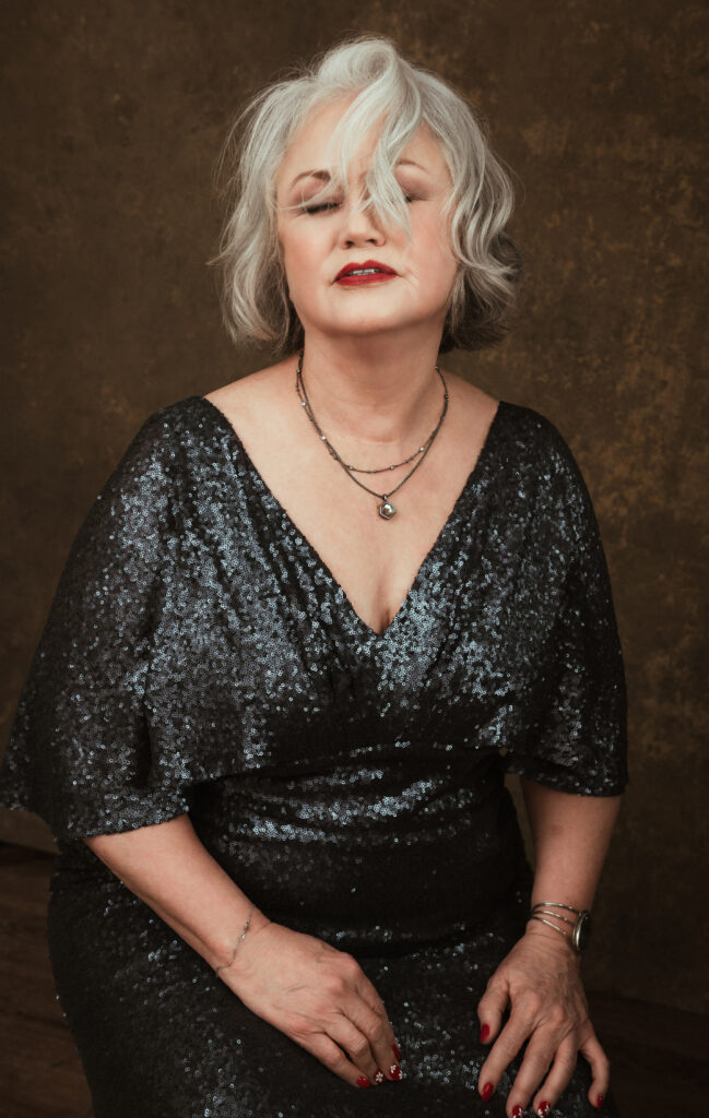 A woman with short gray hair in a dark, sequined dress sits with her eyes closed, wearing layered necklaces and red lipstick against a brown background. Tanya Eivin Photography