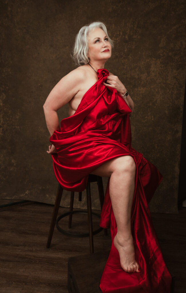 A woman with gray hair sits on a stool, partially draped in a red satin fabric, with a neutral brown background behind her. Tanya Eivin Photography
