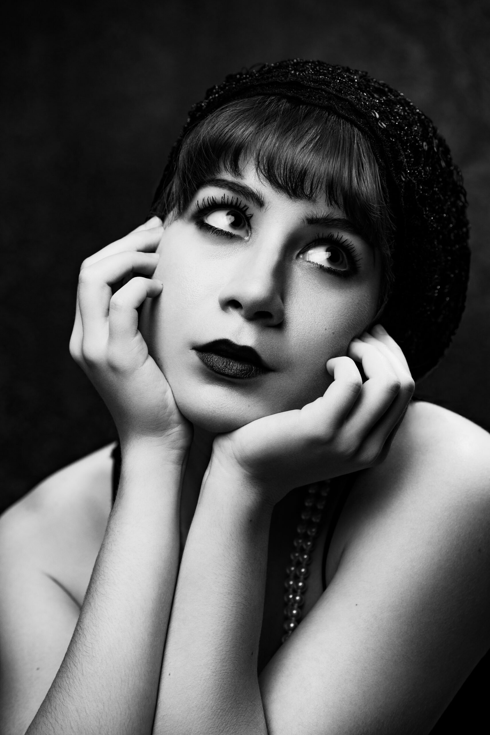 Black and white portrait of a person with short hair, wearing a beaded headpiece and necklace, looking upward while resting their face on their hands. Tanya Eivin Photography