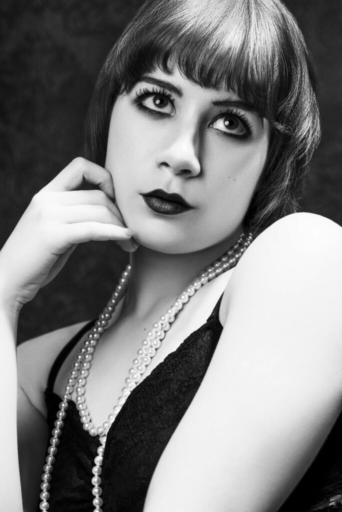 Black and white portrait of a woman with short hair, dark lipstick, dramatic eye makeup, wearing a pearl necklace and a black top, posing with one hand near her face. Tanya Eivin Photography