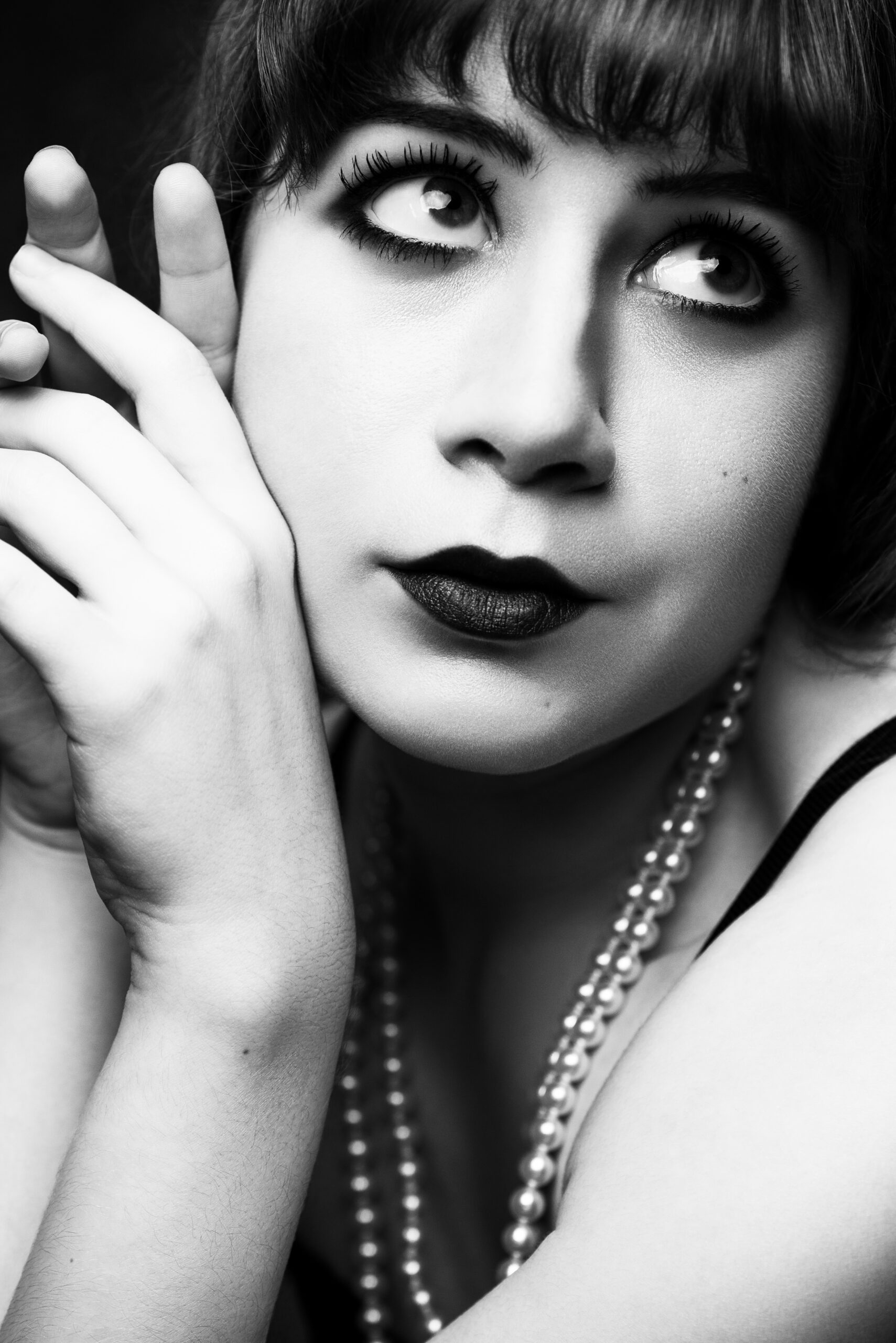 Black and white portrait of a person with dark lipstick, heavy eye makeup, short hair, and a pearl necklace, gazing upward with hands near their face. Tanya Eivin Photography