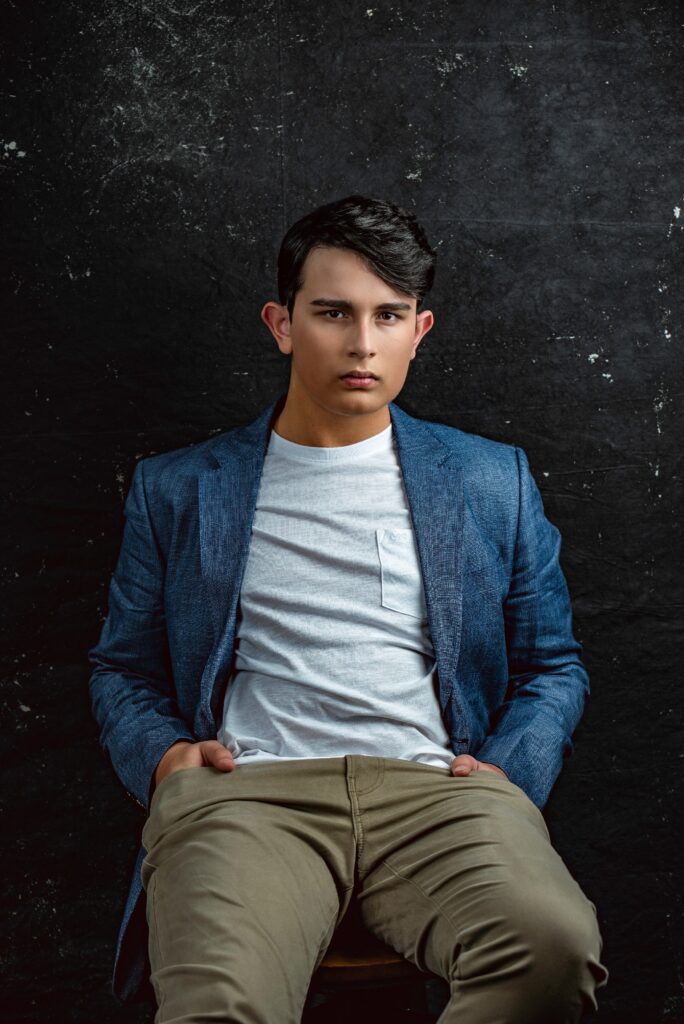 A young man in a blue blazer and light t-shirt sits on a chair against a dark textured background, looking directly at the camera. actor headshots Austin TX