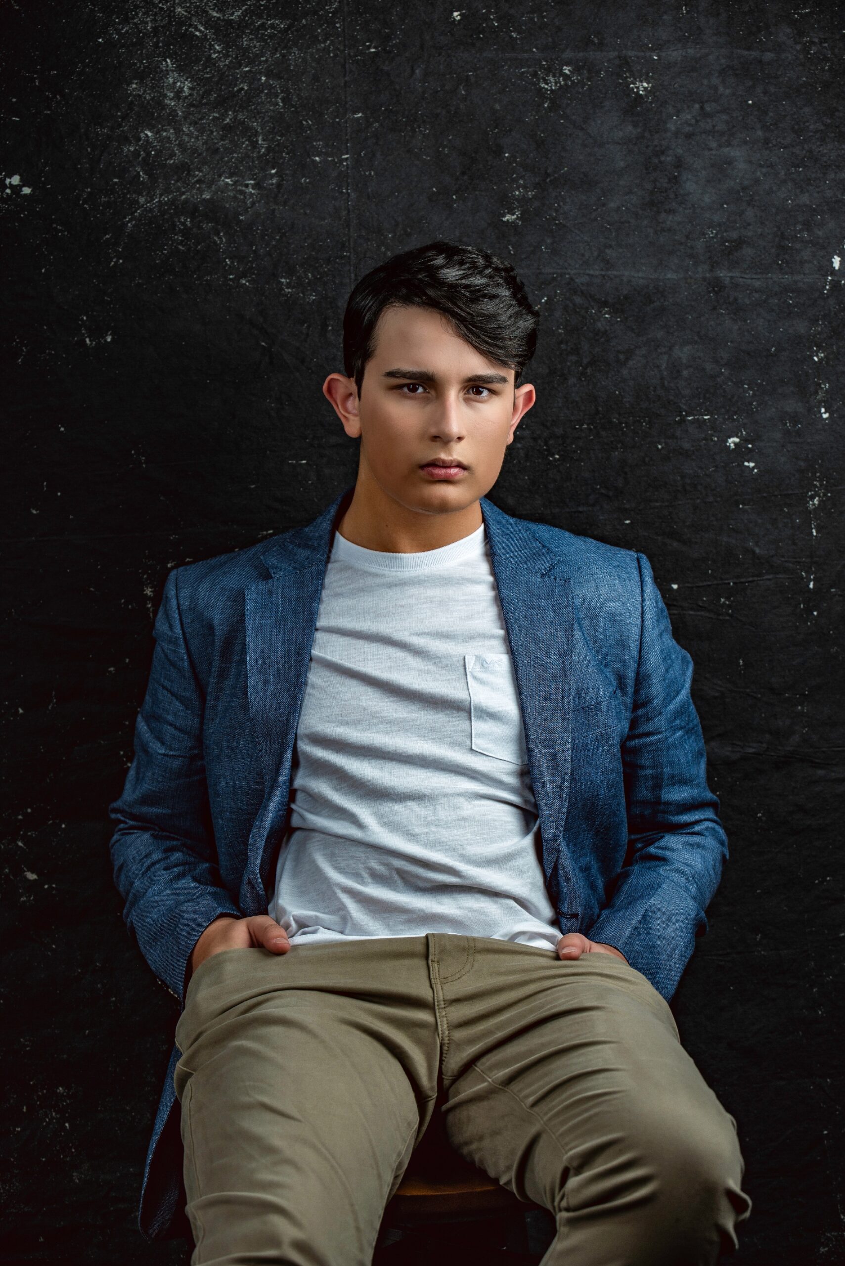 A young man in a blue blazer and light t-shirt sits on a chair against a dark textured background, looking directly at the camera. actor headshots Austin TX