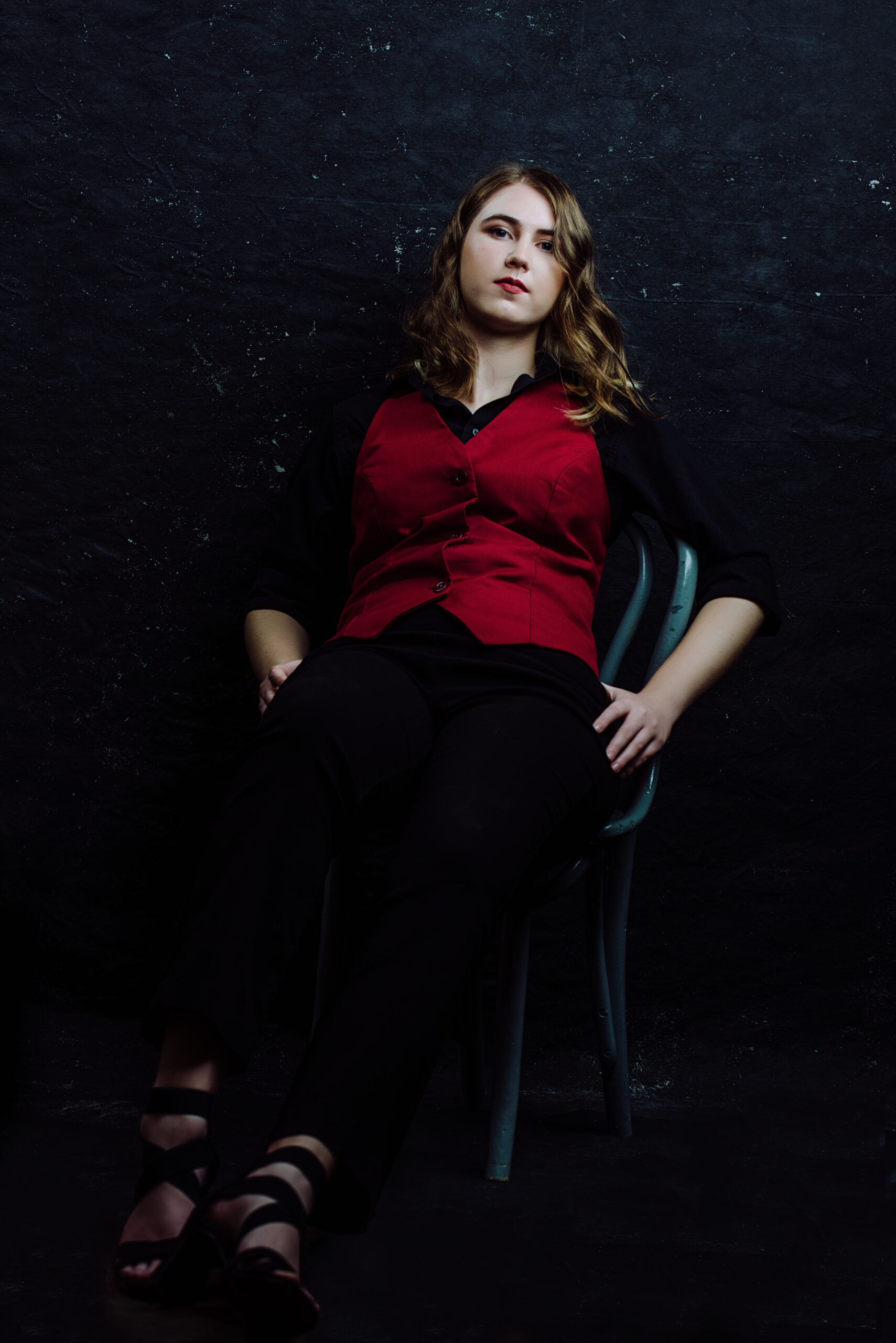 A person with wavy hair, wearing a red vest and black pants, sits on a chair against a dark background, looking straight ahead with a neutral expression. Tanya Eivin Photography
