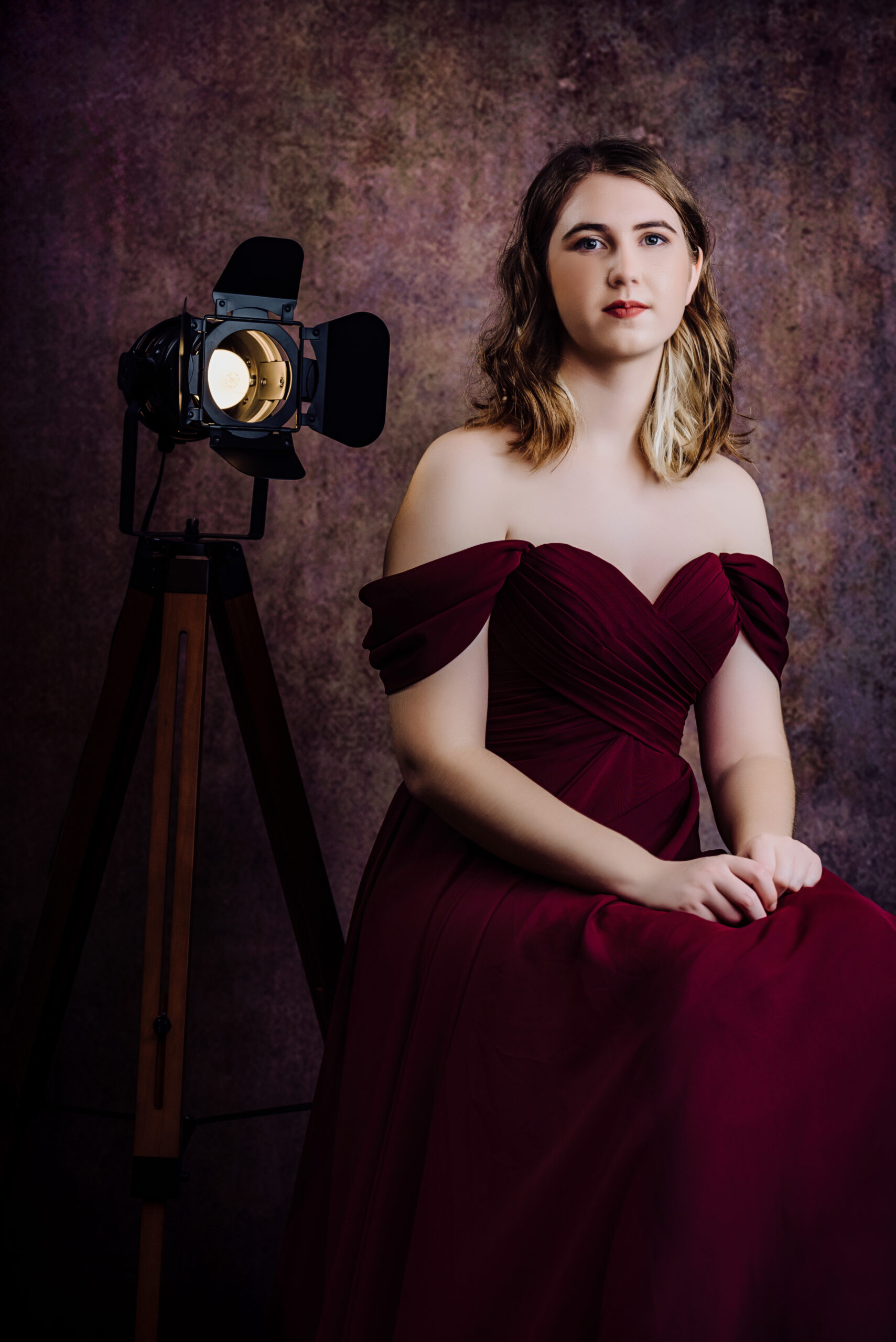 A woman in a burgundy off-shoulder gown sits with her hands folded, next to a studio light on a stand, against a textured backdrop. Tanya Eivin Photography
