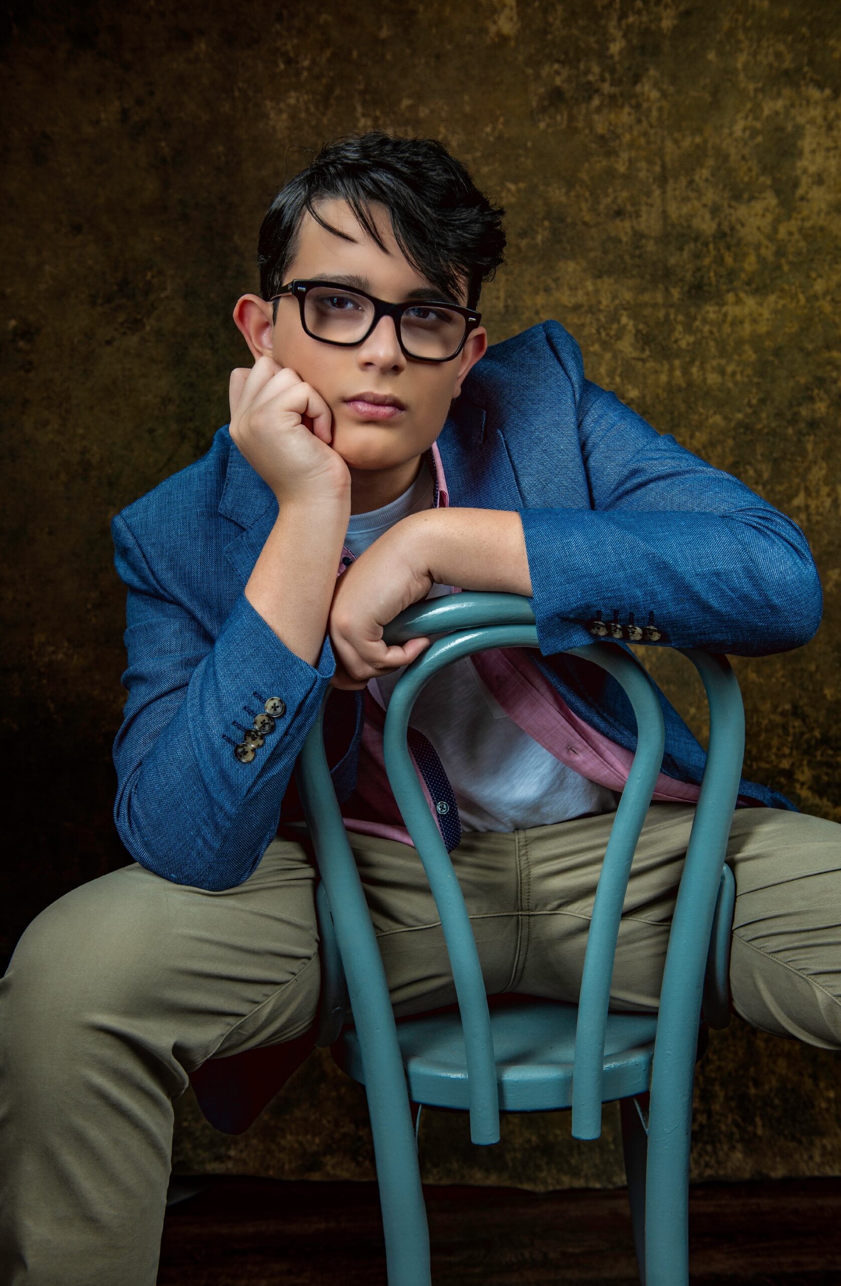 A person with short dark hair, glasses, and a blue blazer sits backward on a blue chair, resting their chin on their hand against a mottled brown background. Tanya Eivin Photography