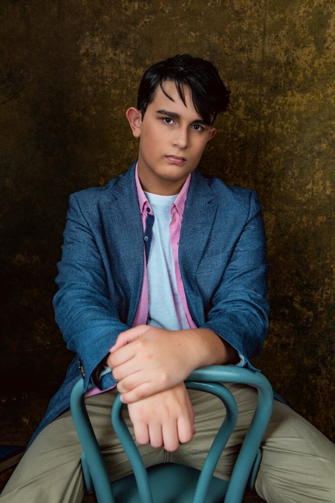A young person with dark hair sits on a blue chair, wearing a blue blazer, pink shirt, and tan pants, facing the camera against a textured brown background. Tanya Eivin Photography
