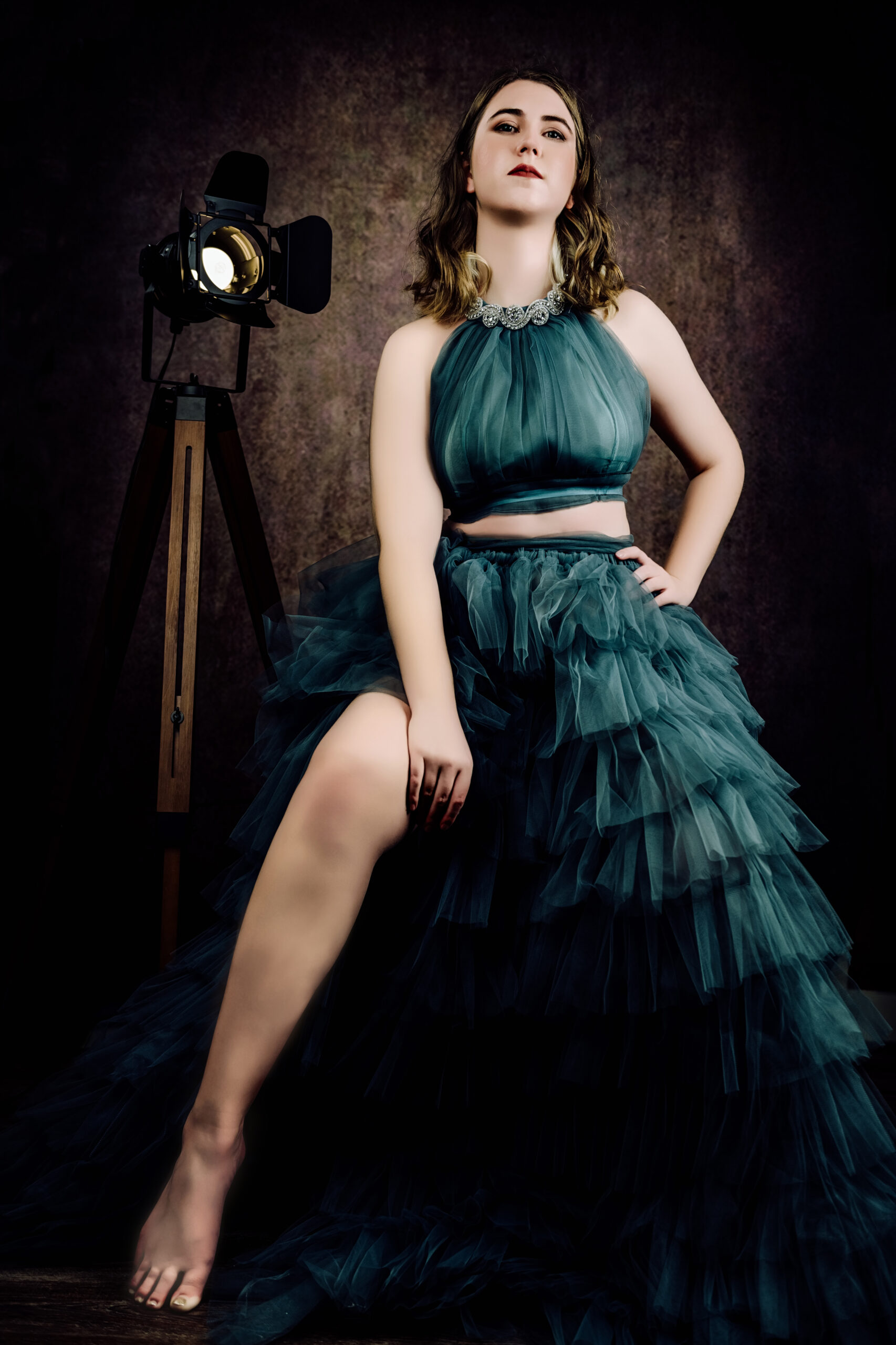 A woman in a dark teal ruffled gown poses confidently with one leg raised, in front of a studio light and a textured brown backdrop. Tanya Eivin Photography