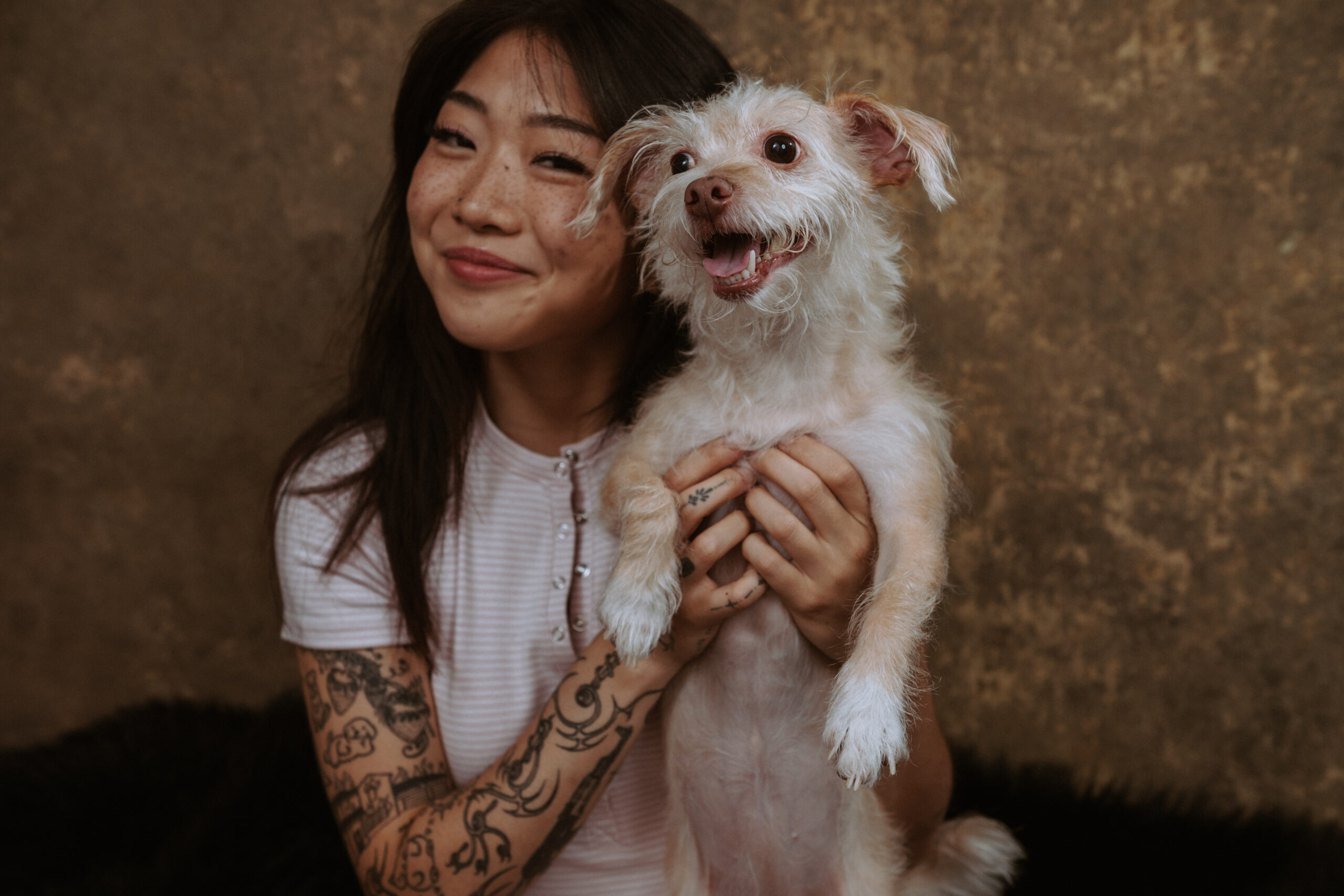 A woman with tattoos smiles while holding a small, scruffy white dog in front of a textured brown background. Tanya Eivin Photography