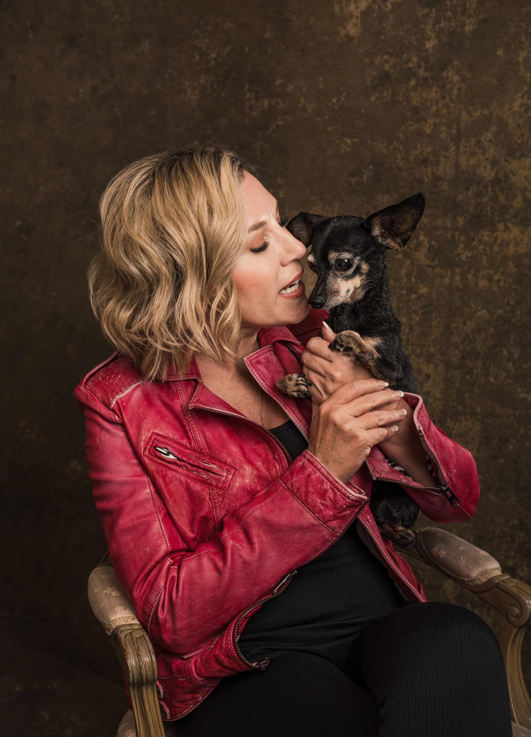 A woman with blonde hair in a red jacket sits on a chair, holding and looking at a small black and brown dog. Tanya Eivin Photography