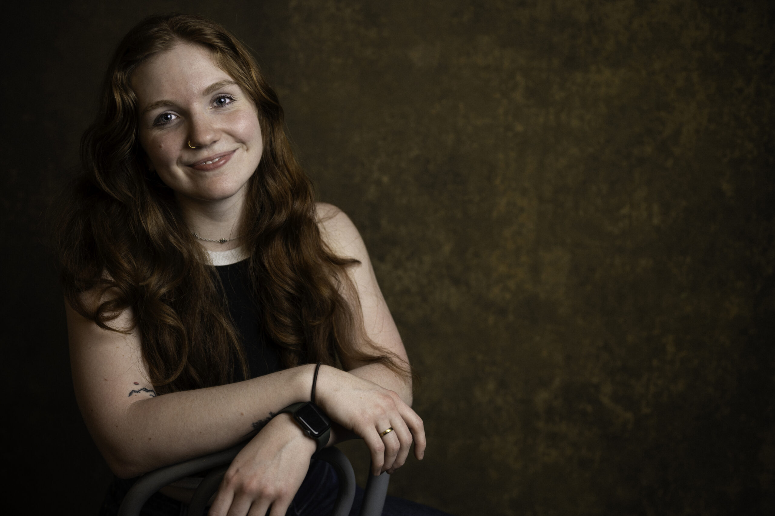 A woman with long red hair sits on a chair, smiling at the camera against a brown textured background. She wears a black sleeveless top and a smartwatch. Tanya Eivin Photography