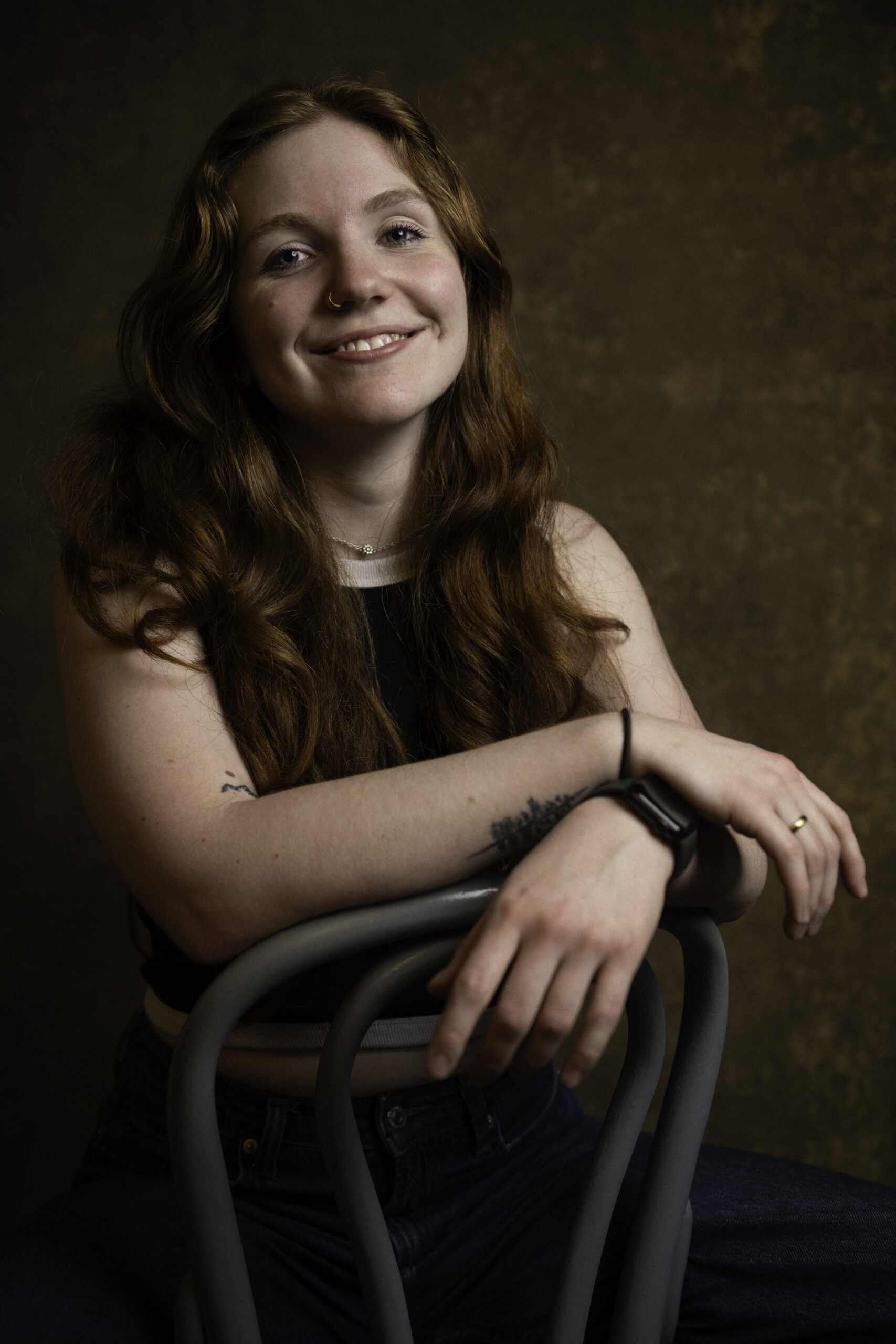 A woman with long red hair sits on a chair backward, smiling at the camera against a brown textured background. She wears a black top, dark jeans, and a smartwatch. Tanya Eivin Photography