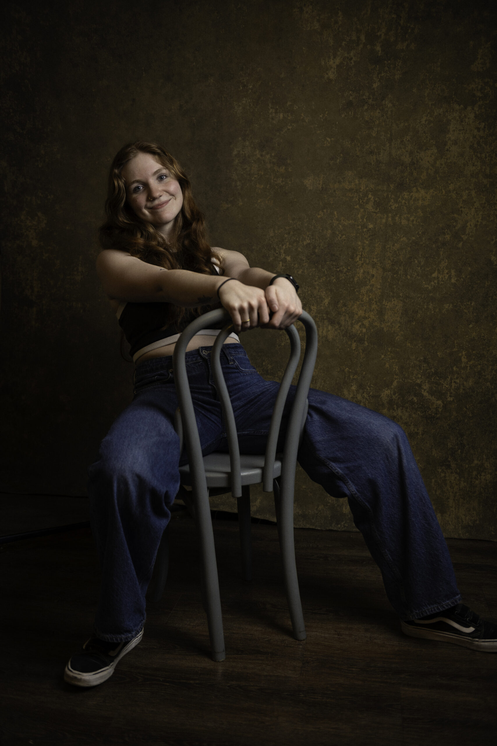 A person with long red hair sits backward on a wooden chair, wearing a black top, blue jeans, and sneakers, against a dark textured background. Tanya Eivin Photography