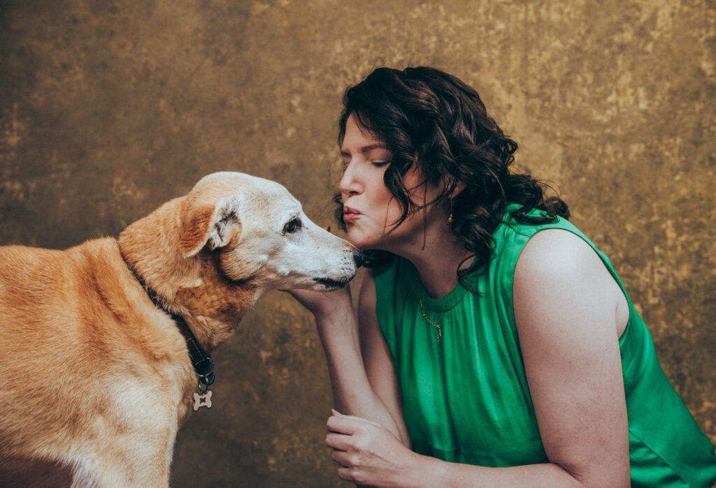 A woman in a green sleeveless top leans in to touch noses with a tan dog wearing a collar, against a textured brown background. Tanya Eivin Photography