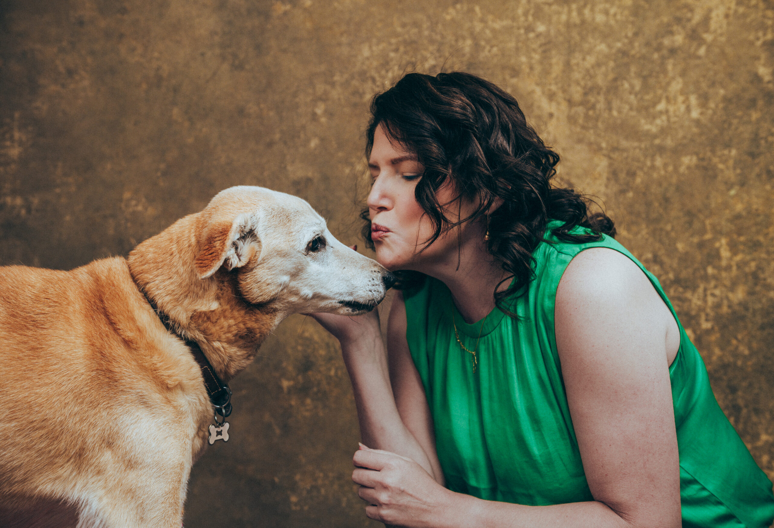 A woman in a green sleeveless top leans in to touch noses with a tan dog wearing a collar, against a textured brown background. Tanya Eivin Photography