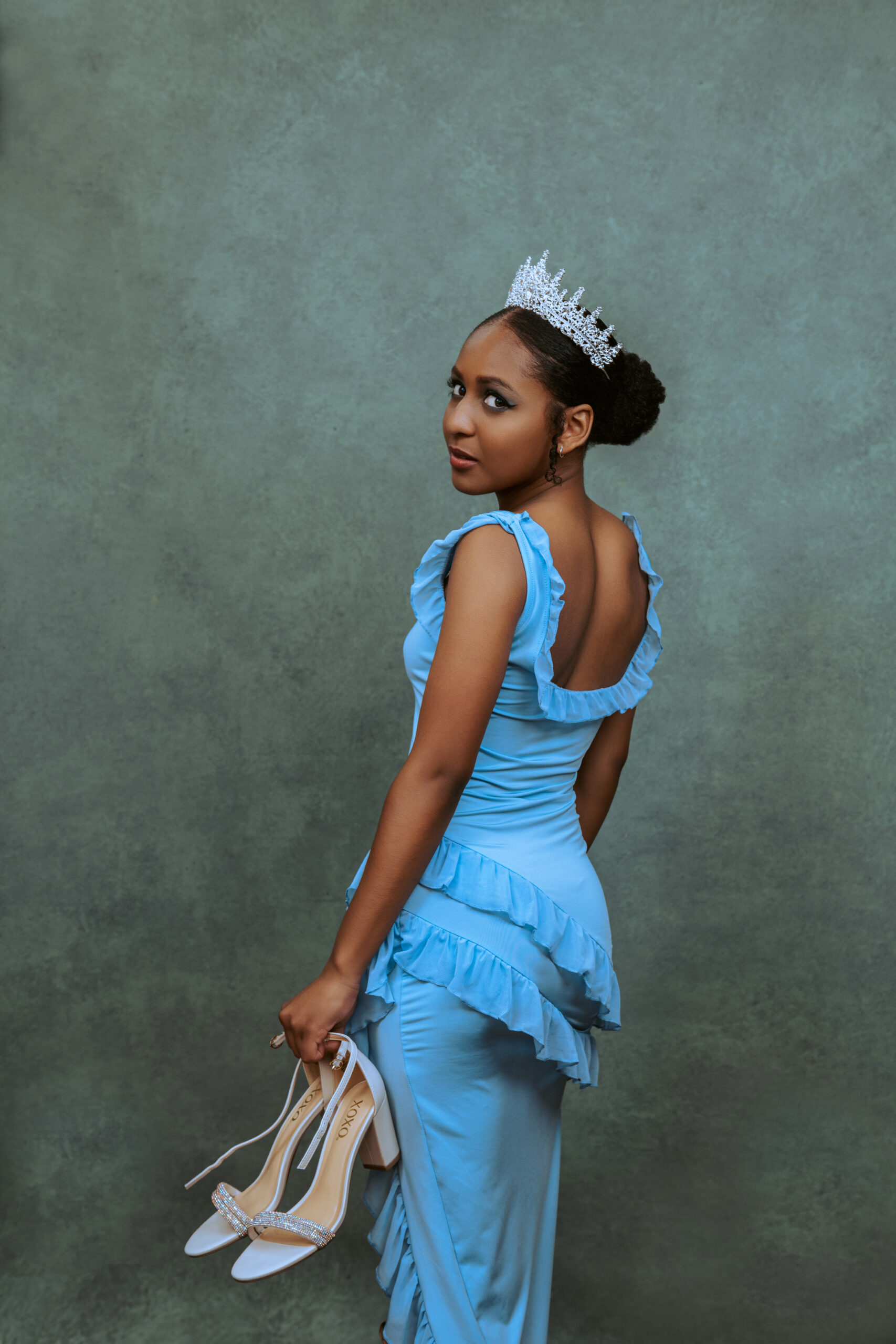 A woman in a light blue ruffled dress and a tiara stands against a green backdrop, holding a pair of high-heeled shoes in her hand. Tanya Eivin Photography