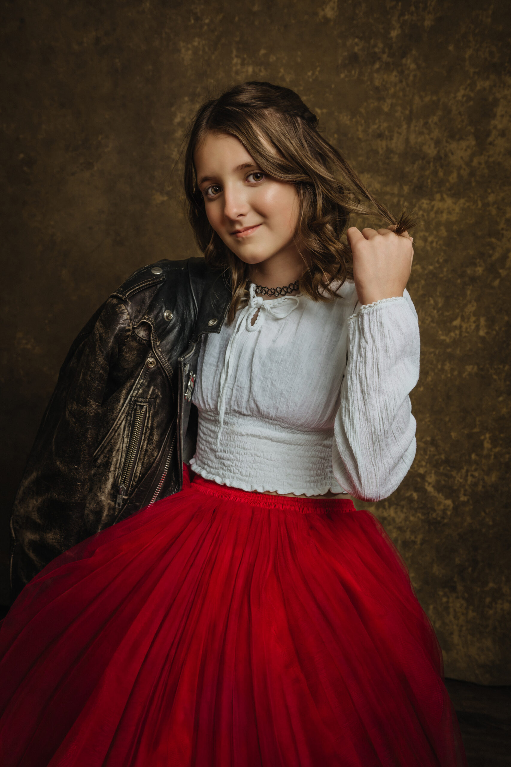A young girl poses in a white blouse, red tulle skirt, and black leather jacket draped over her shoulder, standing in front of a brown textured background. Tanya Eivin Photography