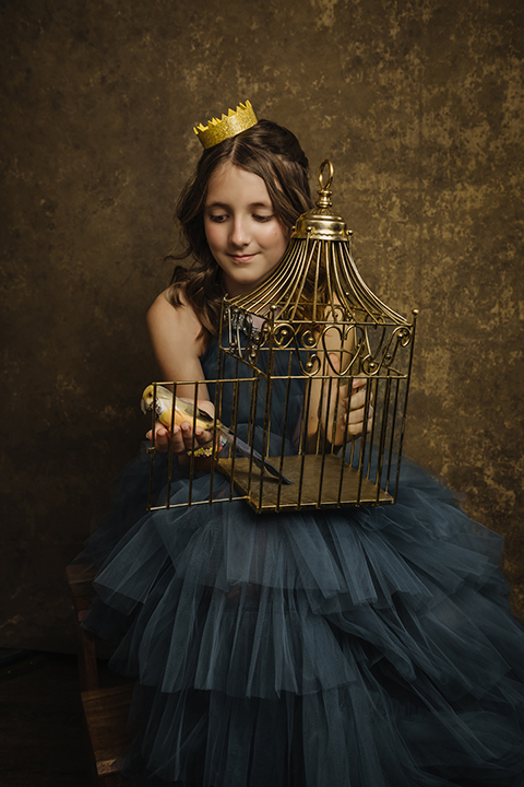 A young girl in a blue tulle dress and yellow crown sits by an open birdcage, gently holding a yellow bird. Tanya Eivin Photography a senior portraits Austin Texas