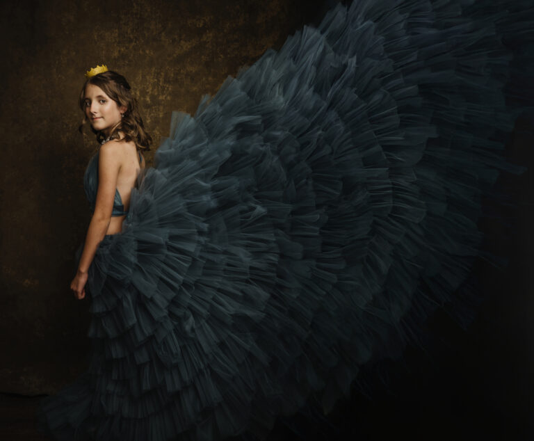 A young teen girl in a beautiful blue tulle gown with an exaggerated, voluminous skirt and a small yellow crown stands against a dark, textured background. Tanya Eivin Photography