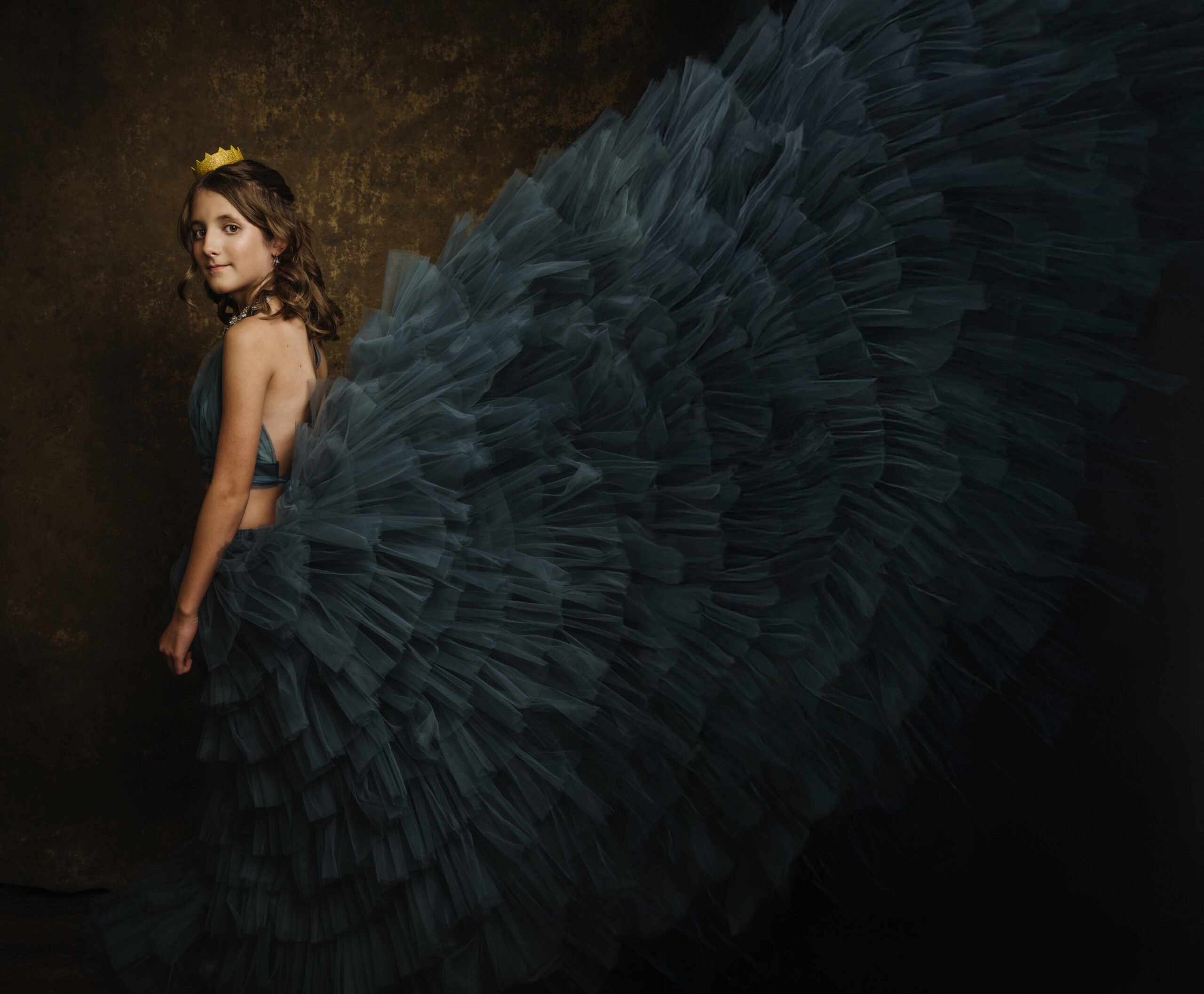 A young teen girl in a beautiful blue tulle gown with an exaggerated, voluminous skirt and a small yellow crown stands against a dark, textured background. Tanya Eivin Photography