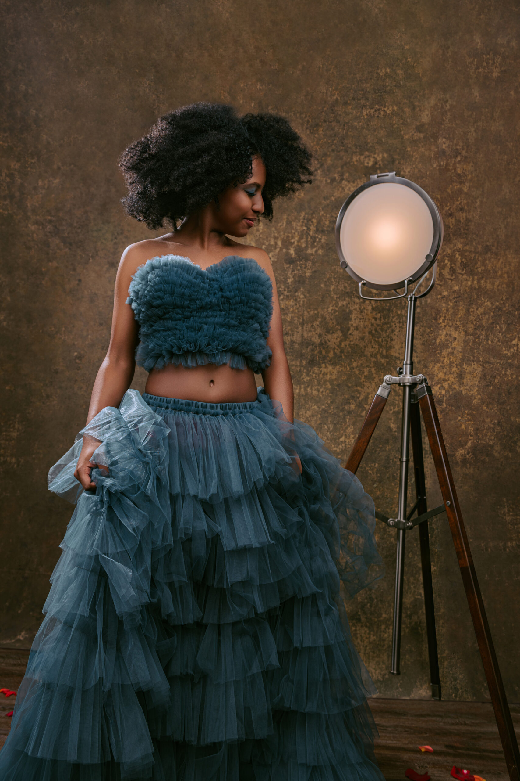 A high school senior in a ruffled teal outfit stands beside a tall tripod lamp, looking down, against a textured brown background with scattered red petals on the floor. Tanya Eivin Photography