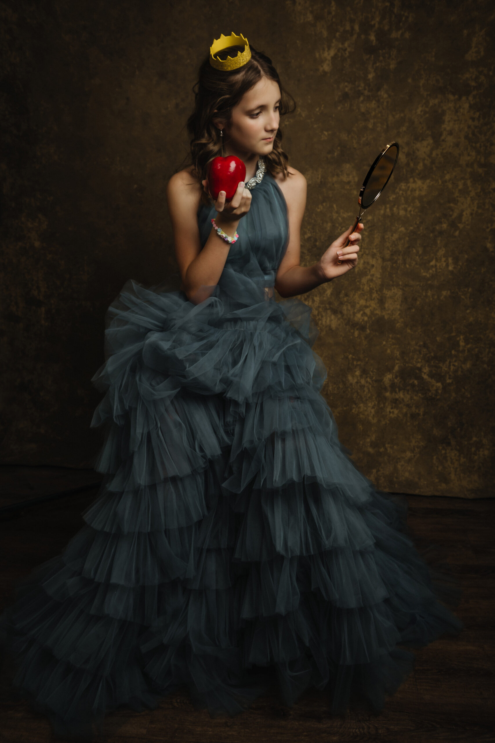A young girl in a ruffled blue gown and yellow crown holds a red apple and a hand mirror, standing against a brown textured background. Tanya Eivin Photography