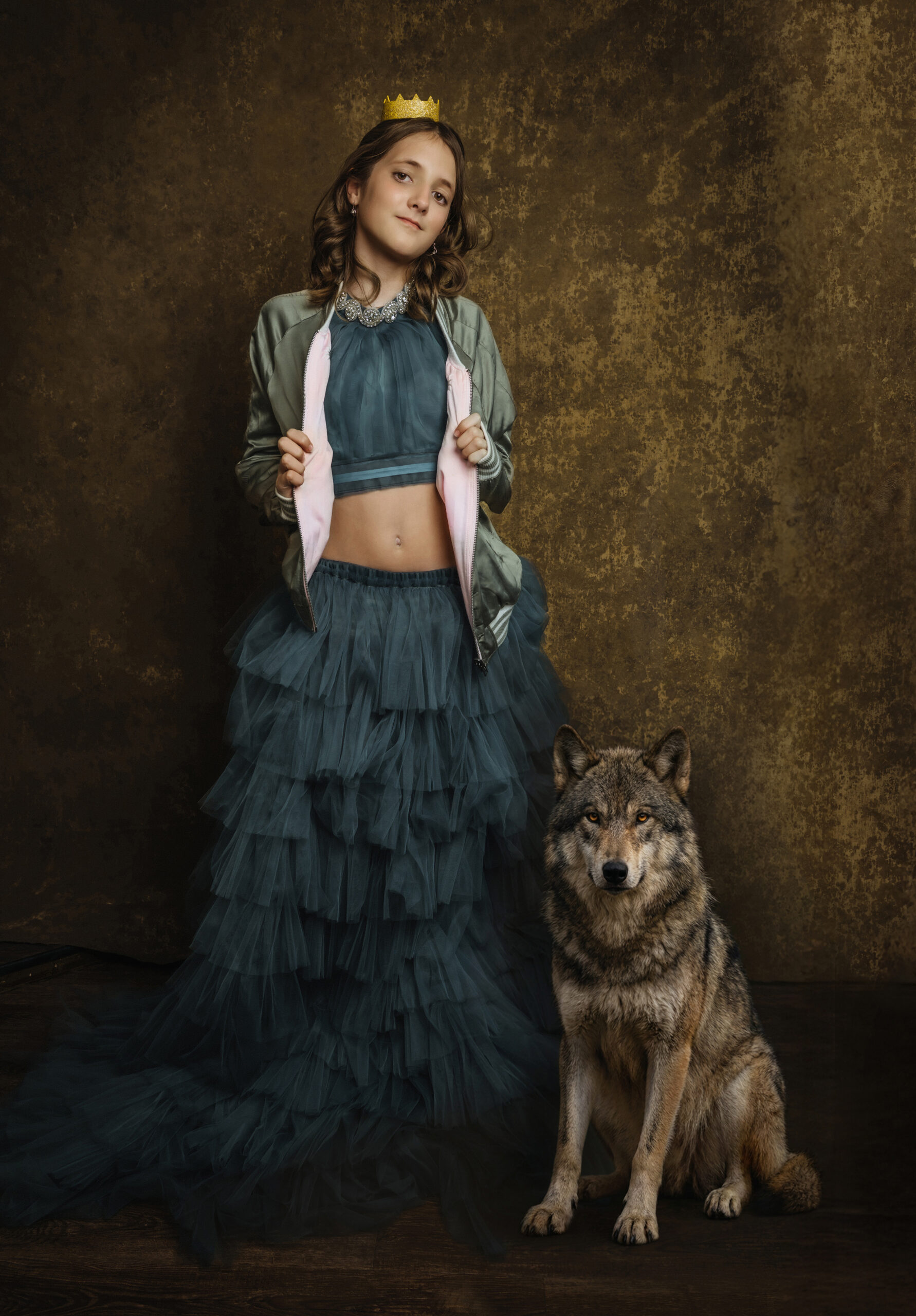 A girl wearing a crown, a blue tulle skirt, and a jacket stands next to a seated wolf against a brown textured backdrop. Tanya Eivin Photography