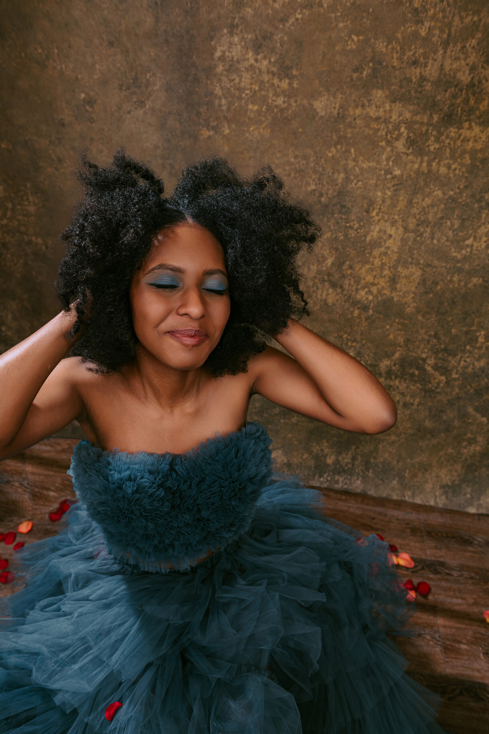 A woman in a blue tulle dress sits with her eyes closed and hands in her hair, against a textured brown background with rose petals on the floor. Tanya Eivin Photography